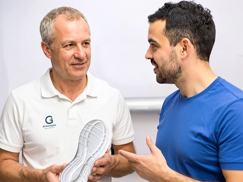 Dois profissionais conversam enquanto analisam um solado branco de calçado. Um deles veste camiseta polo branca com o logotipo da Gi - Inovações para calçados, representando o diálogo técnico e a colaboração no desenvolvimento de componentes para o setor calçadista.